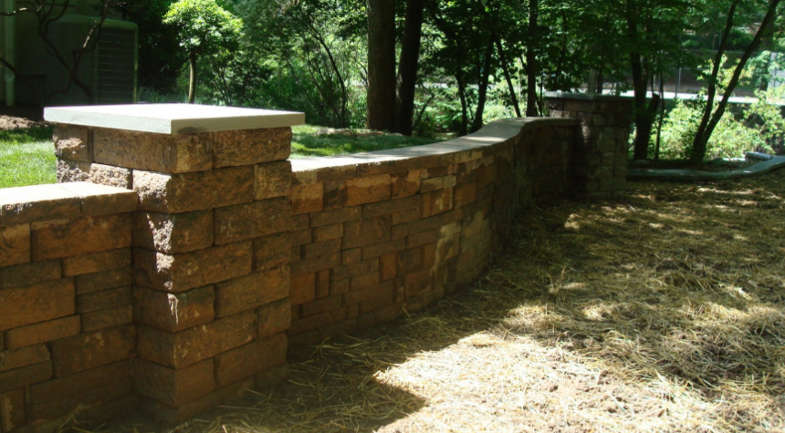 Stone retaining wall with light-colored cap, built in a grassy yard, with trees in the background.