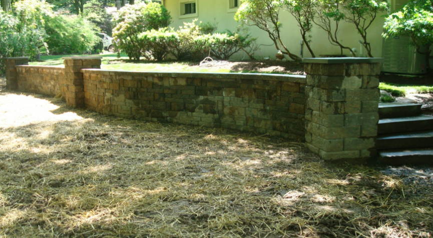 Stone retaining wall in front of a house, with steps leading up. Brown grass in the foreground.