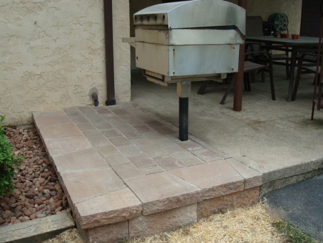 Brick patio with built-in grill, set against a stucco wall. Brown and beige tones dominate the scene.