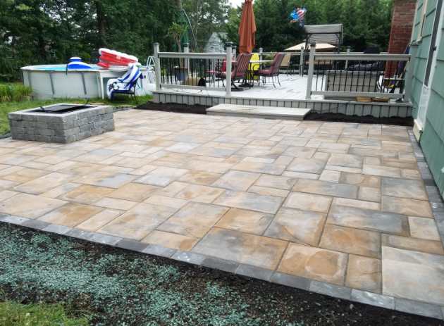 Stone patio with fire pit, deck, and green lawn. Light and dark brown pavers.