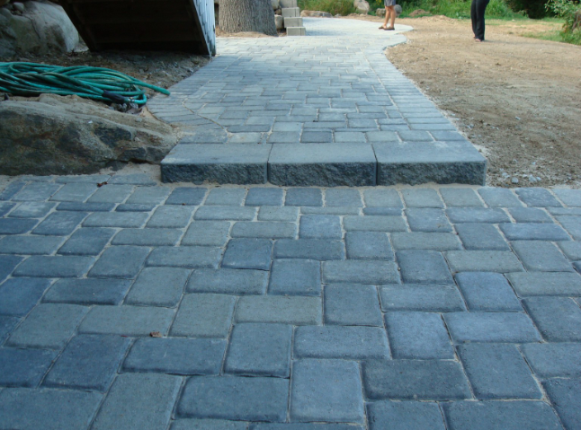 Brick walkway and steps lead towards a person standing on dirt and grass. Blue-grey pavers.