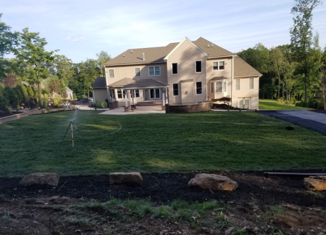 Large beige house with green lawn, patio, and black mulch with boulders; a sprinkler is running.