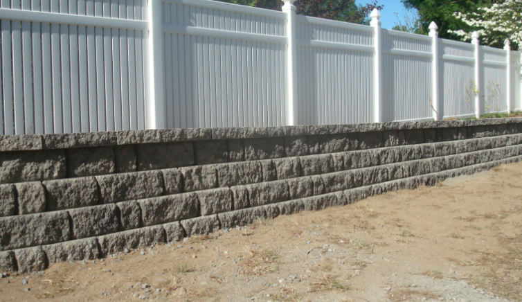 A tiered retaining wall made of gray stone supports a white fence on a sunny day.