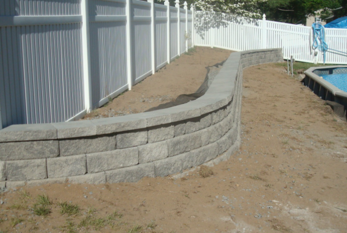 A curved retaining wall made of gray blocks next to a white fence. Sand and grass visible.