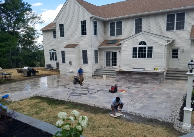 Workers installing a stone patio near a large, white house with multiple windows.