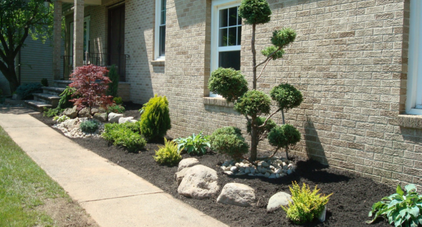 Landscaped front yard with walkway, bushes, rocks, and topiary tree against a brick house.