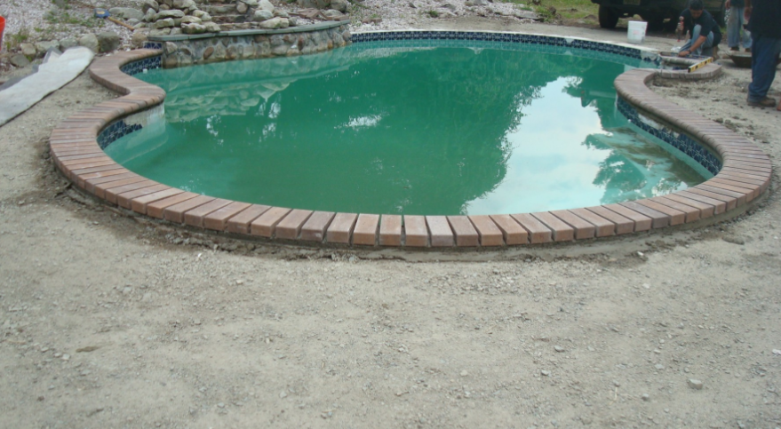 Pool under construction with brick coping and murky green water.