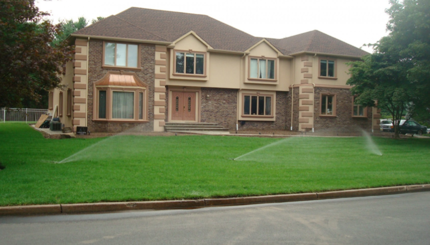 Two-story house with green lawn, brick and beige facade. Sprinklers water the grass.