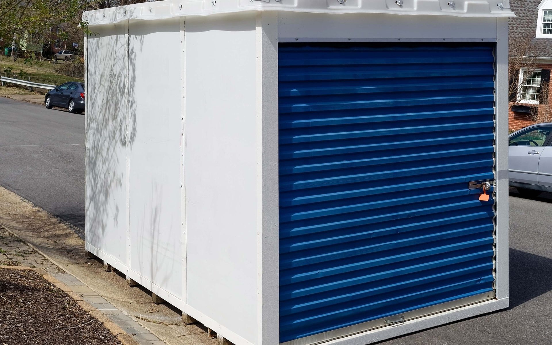 Small white storage shed with a blue roll-up door beside a roadside.