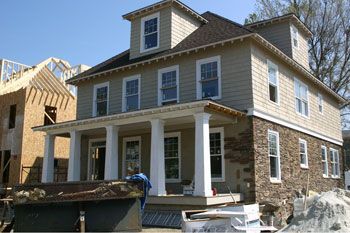 Two-story house under construction with stone and beige siding, white columns, blue tarp, sunny day.