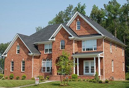 Red brick two-story house with a dark roof and white trim. Green lawn and trees in the background.