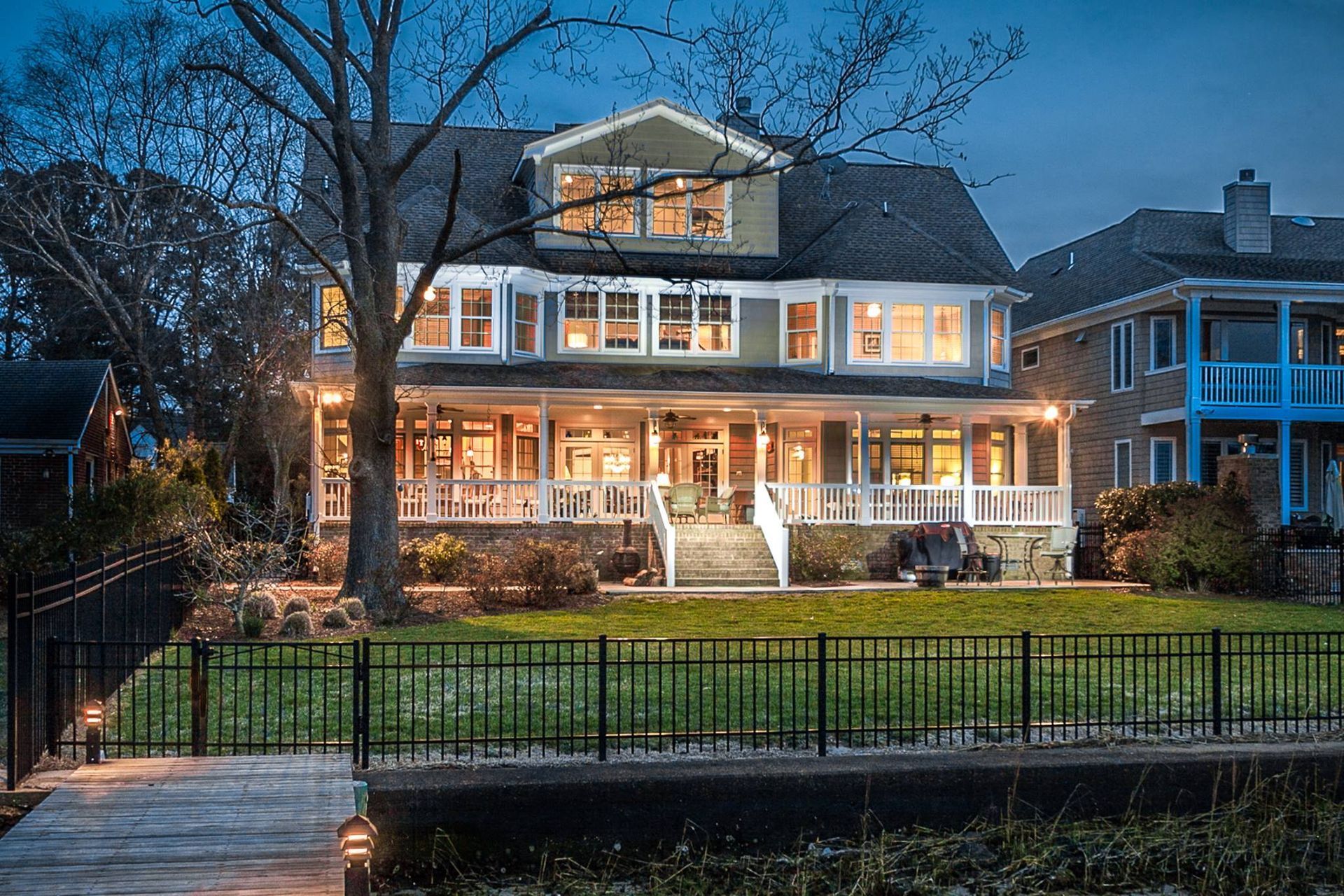 Two-story house at dusk, with lights on inside and on the wrap-around porch, next to a black fence and a waterway.