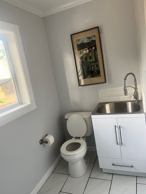 Small bathroom with a toilet, sink, and framed picture. Light gray walls, white cabinets, and a window.