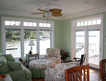 Living room with large windows overlooking water, floral furniture, green walls, and a ceiling fan.