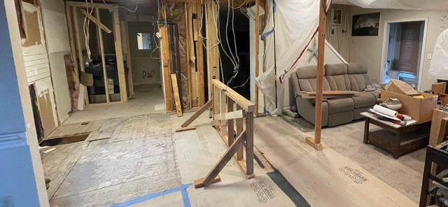 Interior of a room undergoing renovation; exposed wall studs, construction materials, sofa, and boxes.