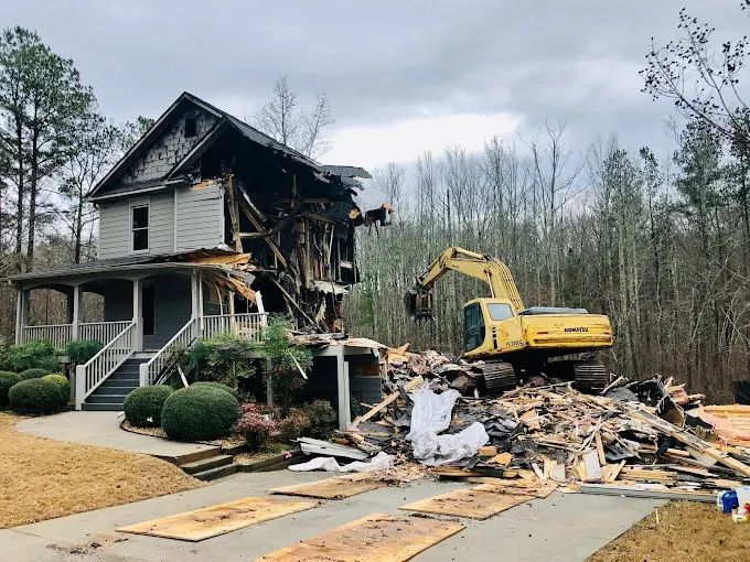 House partially demolished by yellow excavator; debris pile in front. Overcast day.
