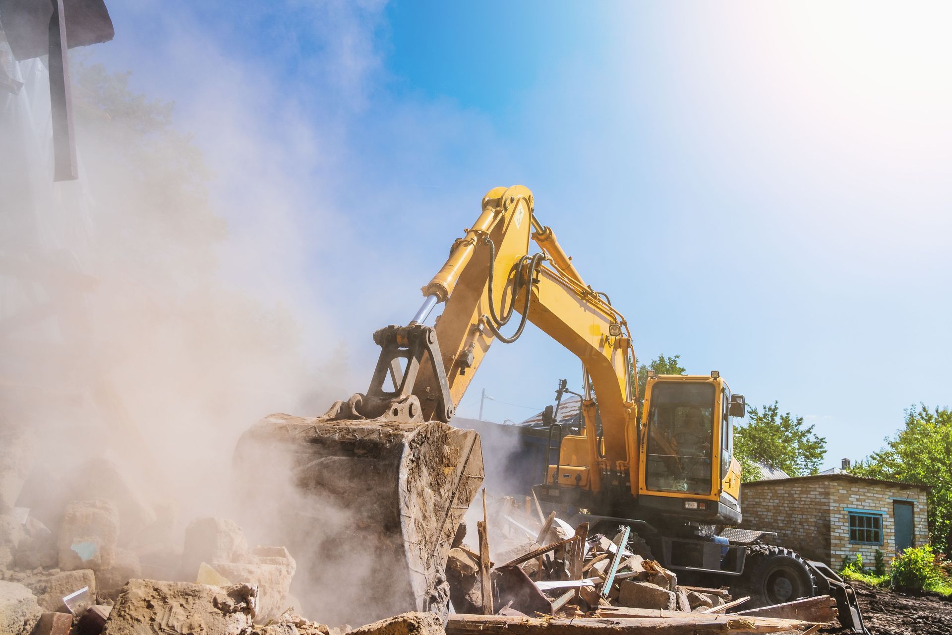 Yellow excavator demolishing a building, generating dust under a blue sky.