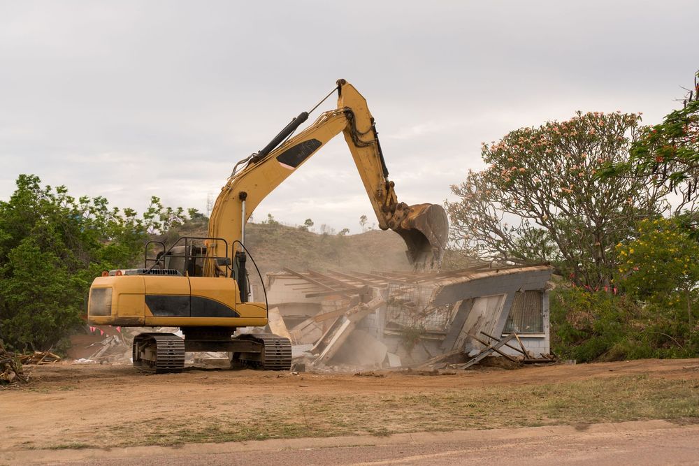 Yellow excavator demolishing a building, debris flying, outdoor setting, cloudy sky.
