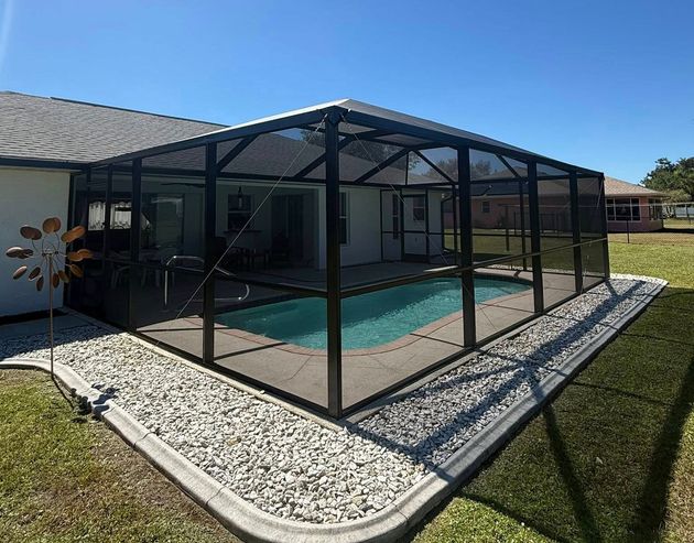 Screened pool enclosure beside a house, surrounded by gravel and lawn under a blue sky