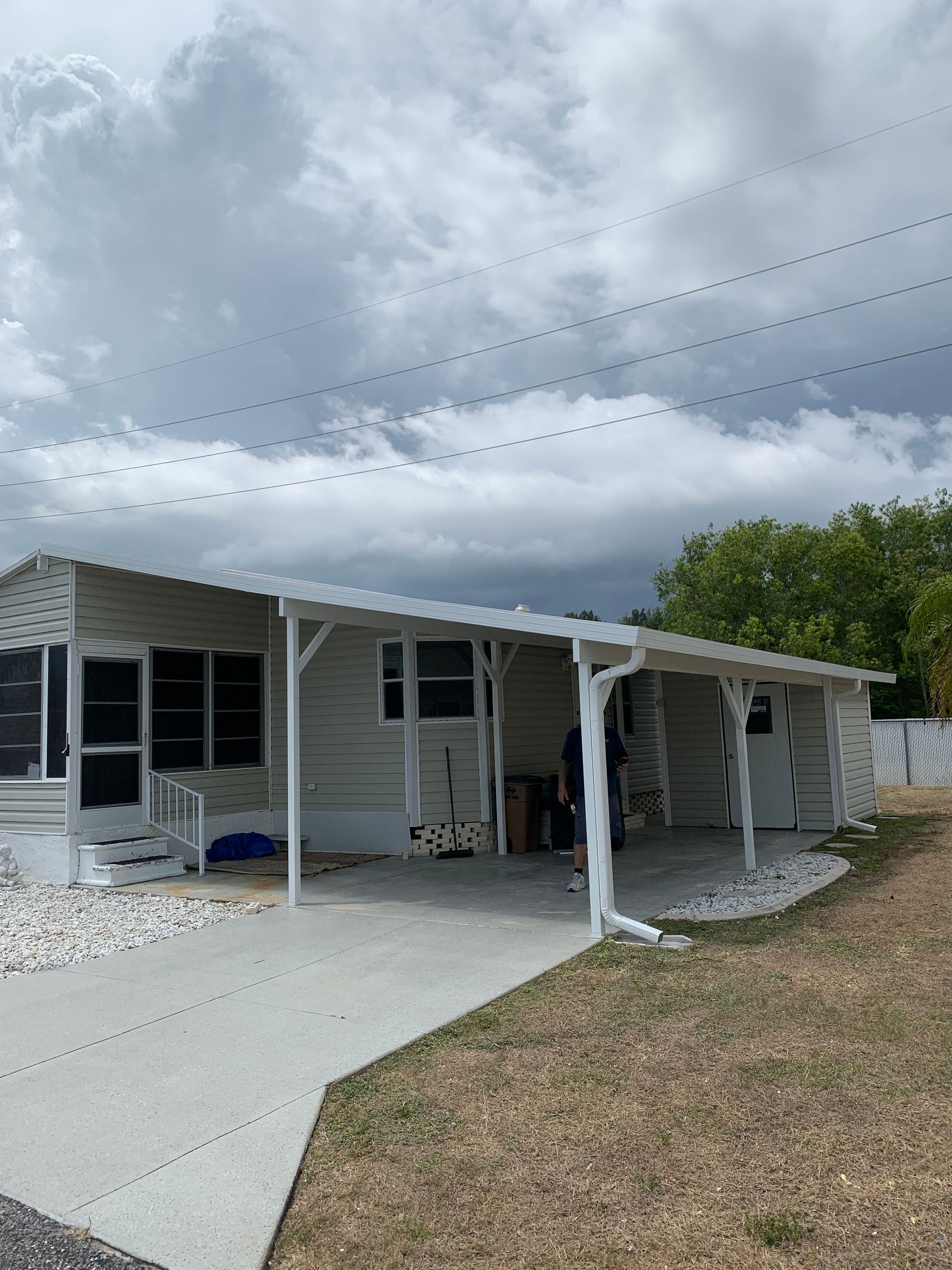 Single-story white house with porch, driveway, and cloudy sky, with cars parked under the carport.