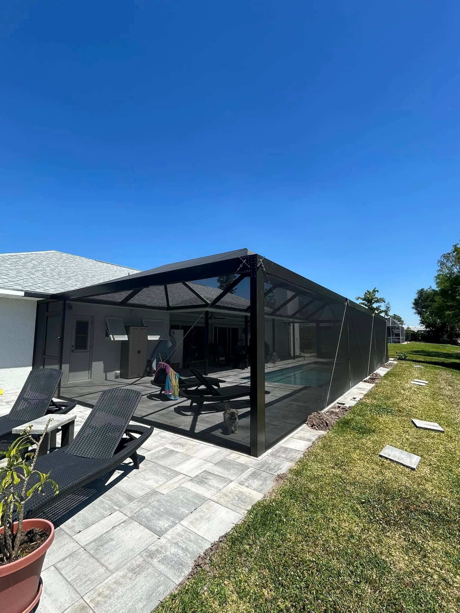 Black screened patio enclosure beside a house, with lounge chairs and a potted plant under a blue sky