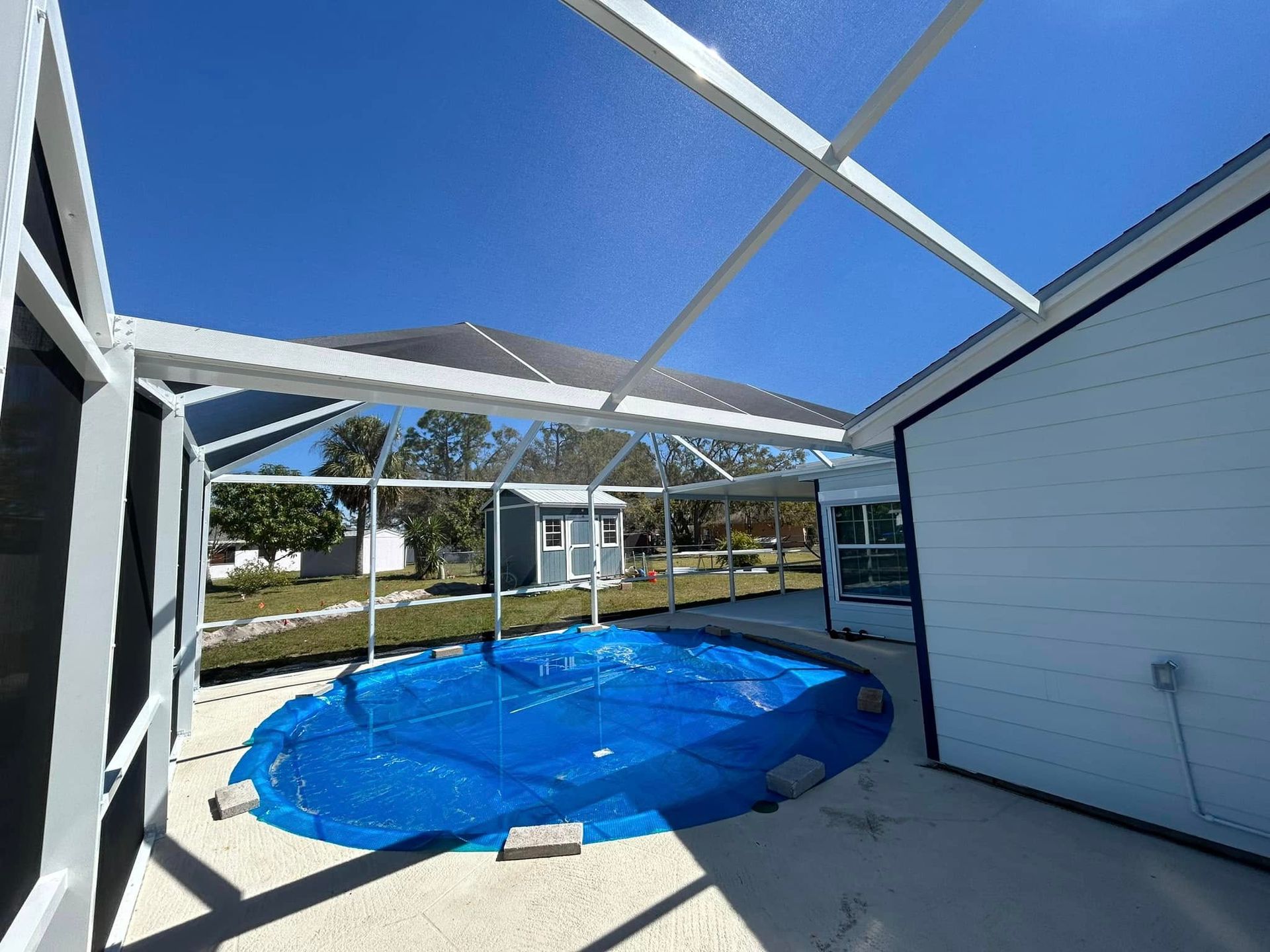 Screened patio with a blue-covered pool and white pergola roof framing a sunny yard