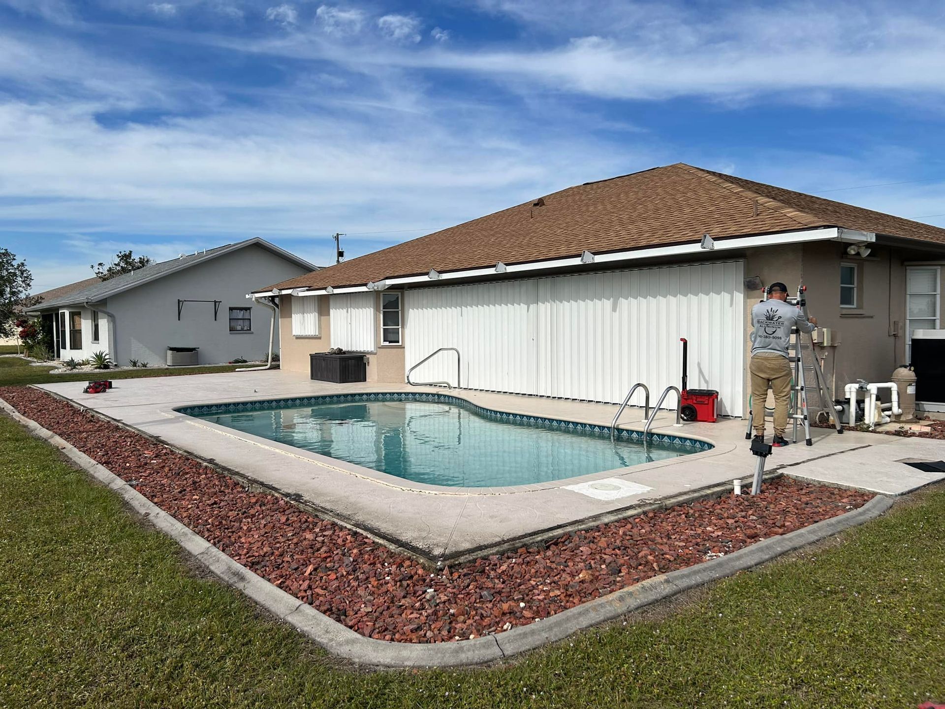 Backyard swimming pool beside a beige house with a red-tiled roof and a person standing near the patio