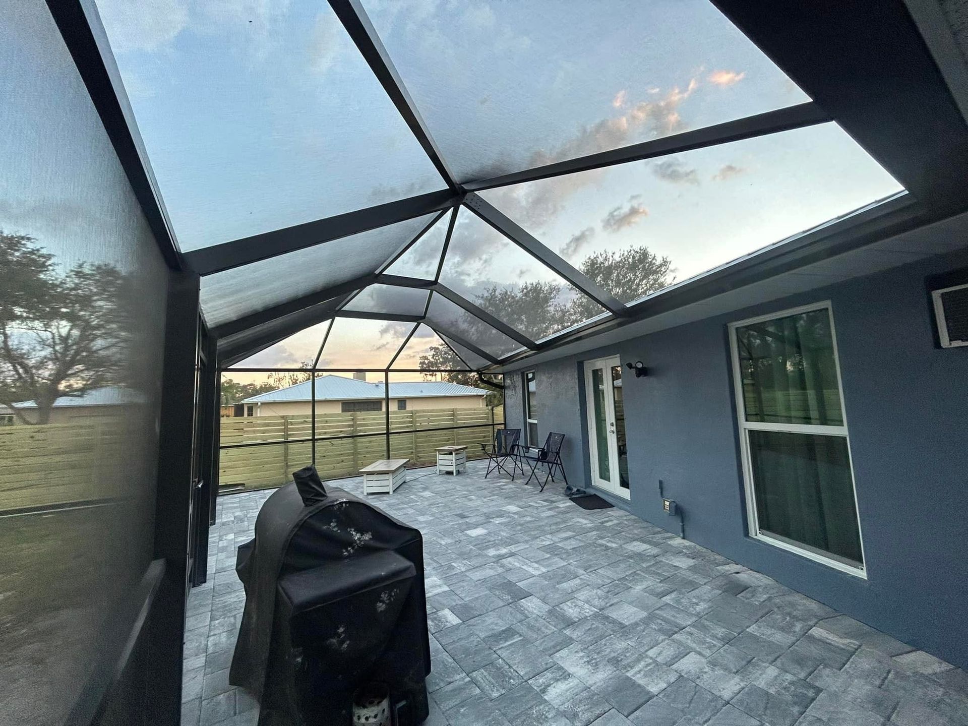 Screened patio with gray pavers, a black grill, and a view of trees and sky beyond the enclosure.