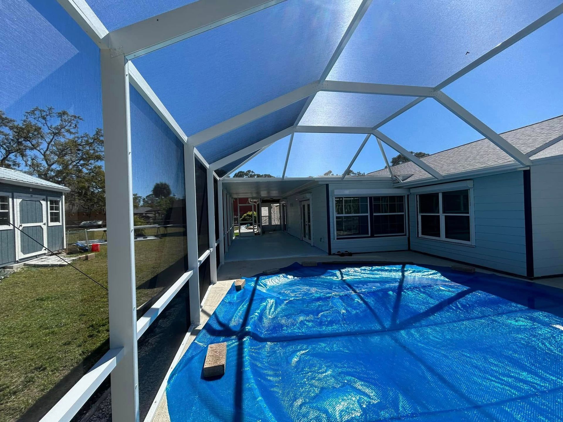 Screened patio with blue tarp floor and white framing beside a house under a clear sky