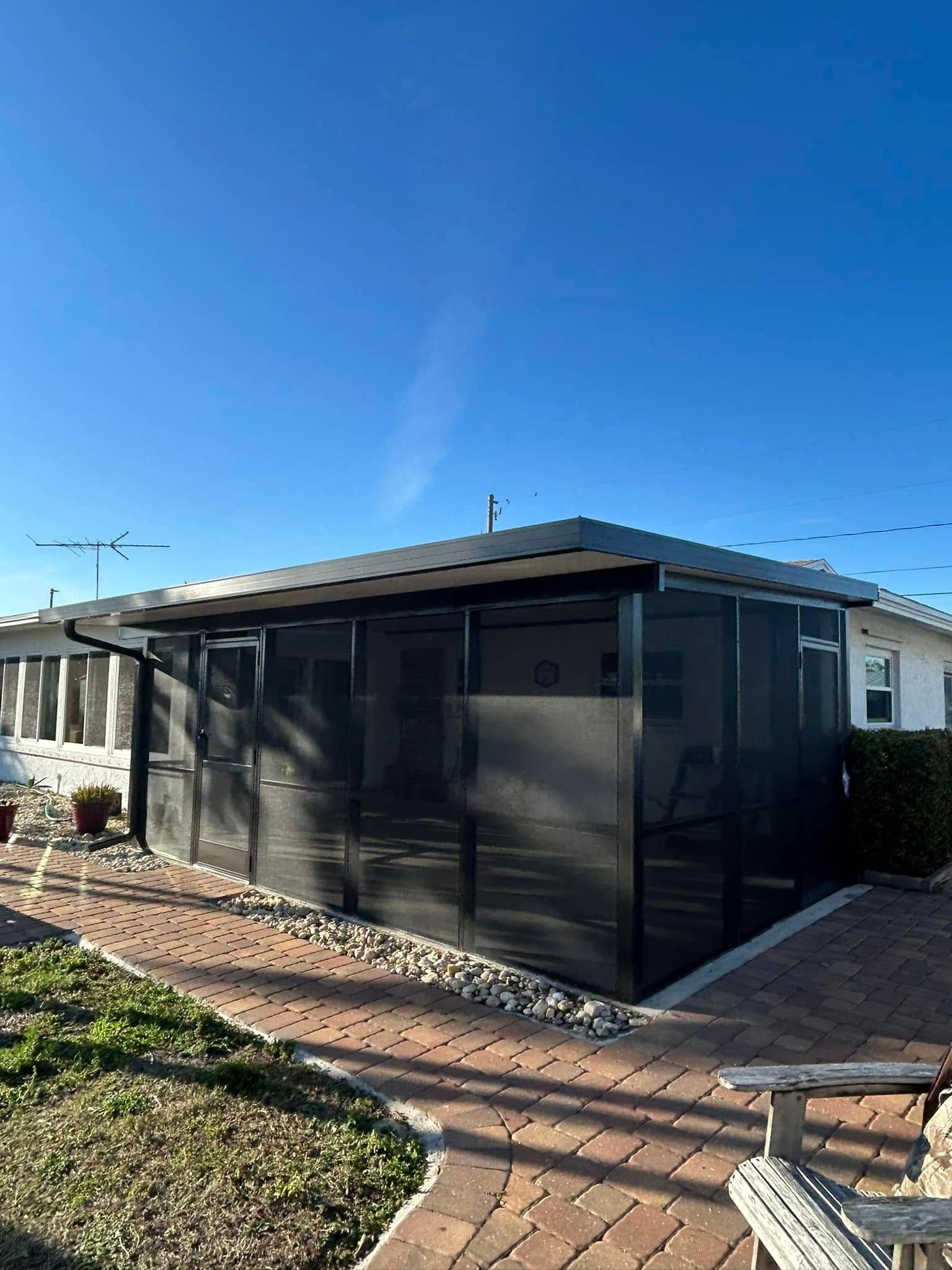 Black screened patio enclosure beside a house, with a paved walkway and bright blue sky.