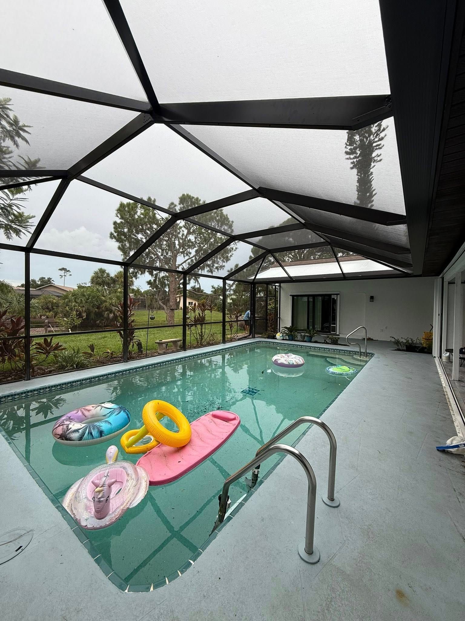 Covered backyard pool with floating rings and a pink mat beside a screened patio.