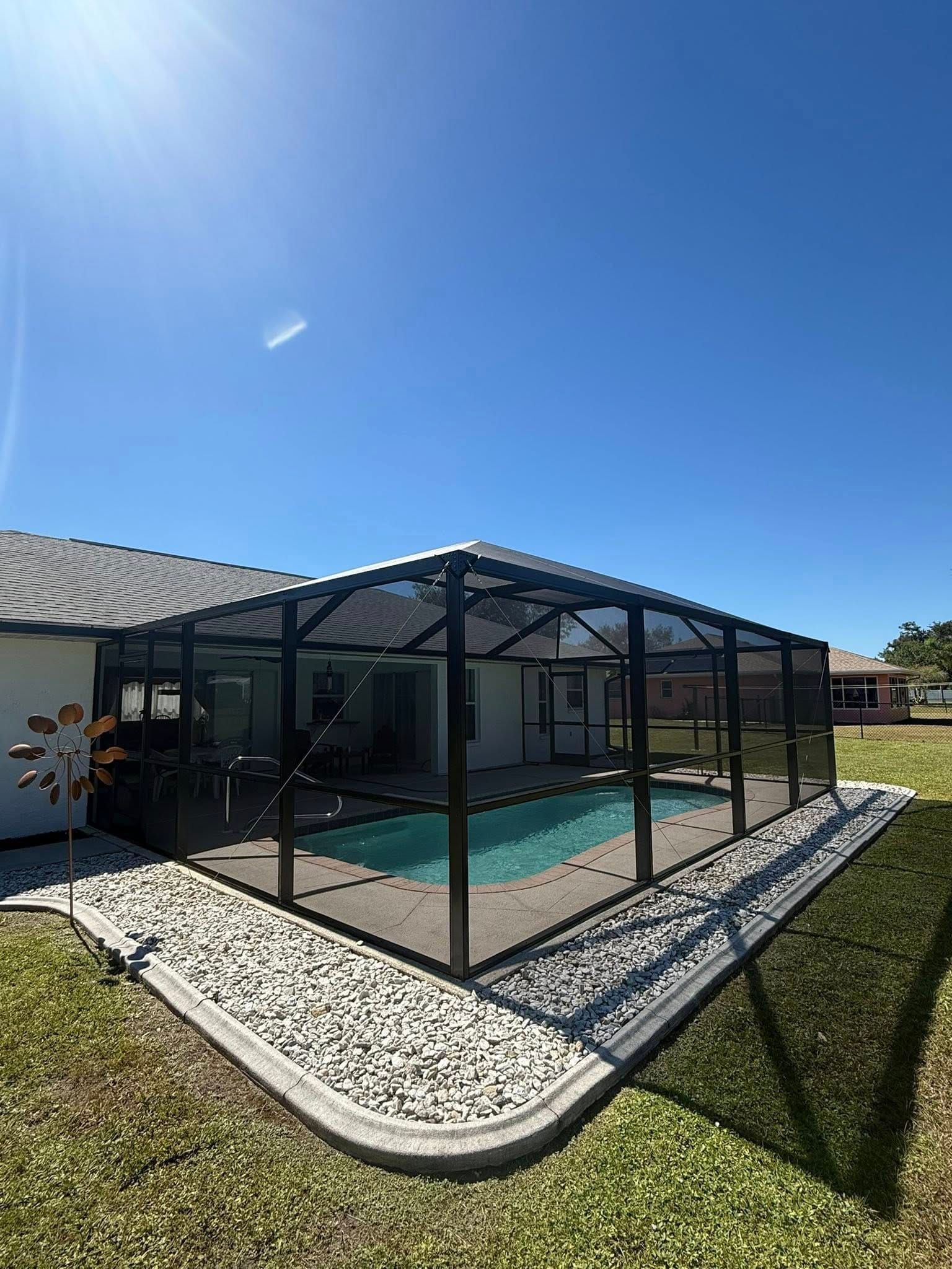 Screened-in backyard pool enclosure beside a house, with blue sky and white rock landscaping
