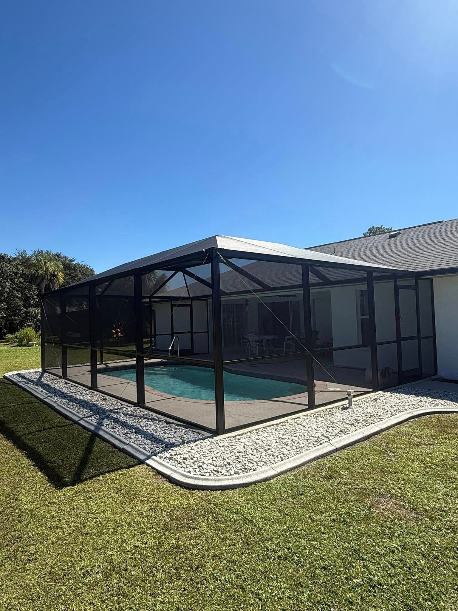 Black screened pool enclosure beside a house on a sunny lawn with blue sky