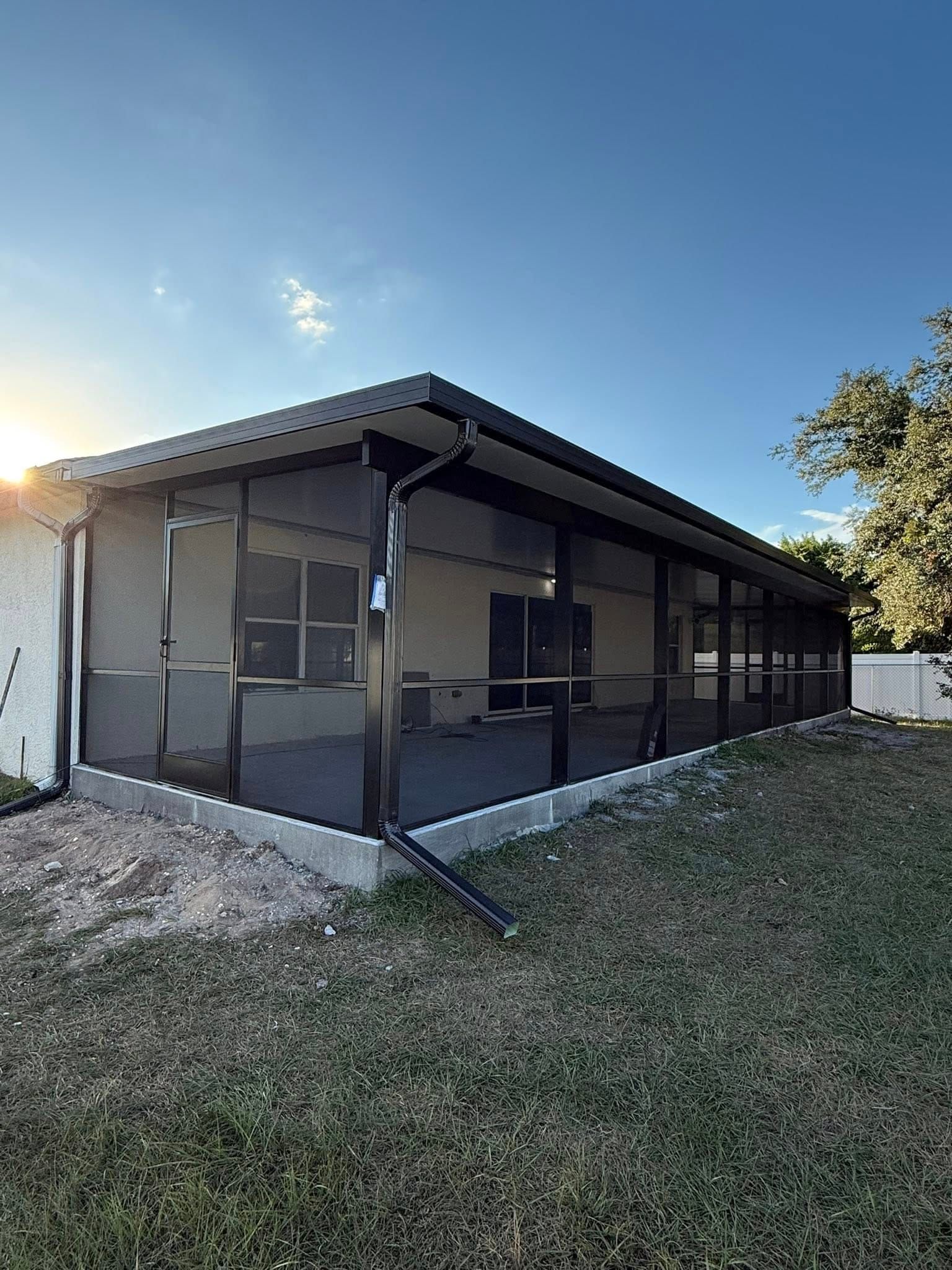 Screened porch on a house with a black roof and mesh walls, viewed from a grassy yard at sunset