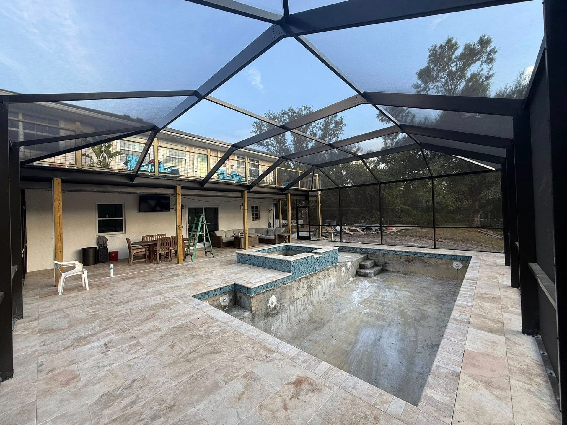 Screened patio with a large empty pool, stone deck, and house seating area under a black mesh enclosure