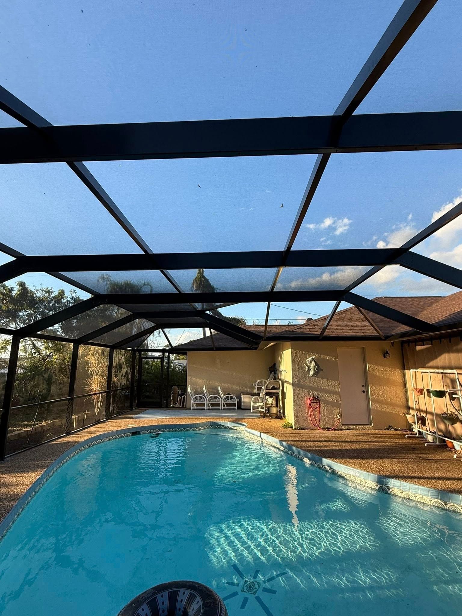 Backyard swimming pool under a screened enclosure, with a beige house and patio in the background.