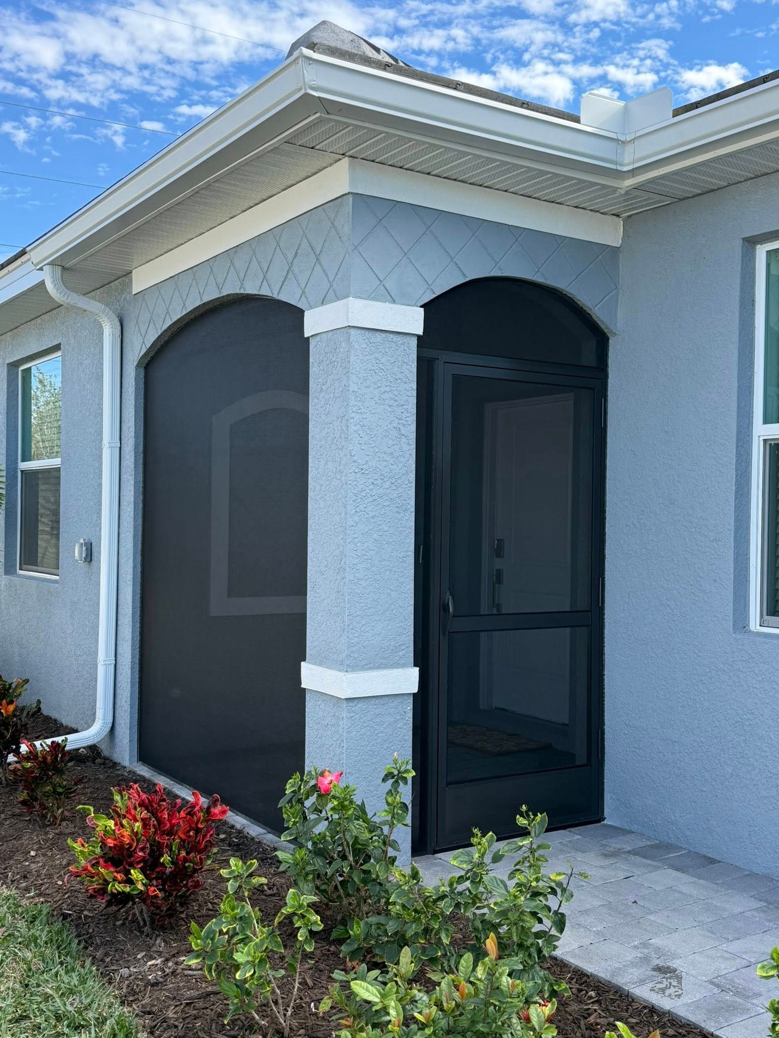 Covered front entry with arched porch, gray stucco walls, dark screen door, and flower bed under blue sky