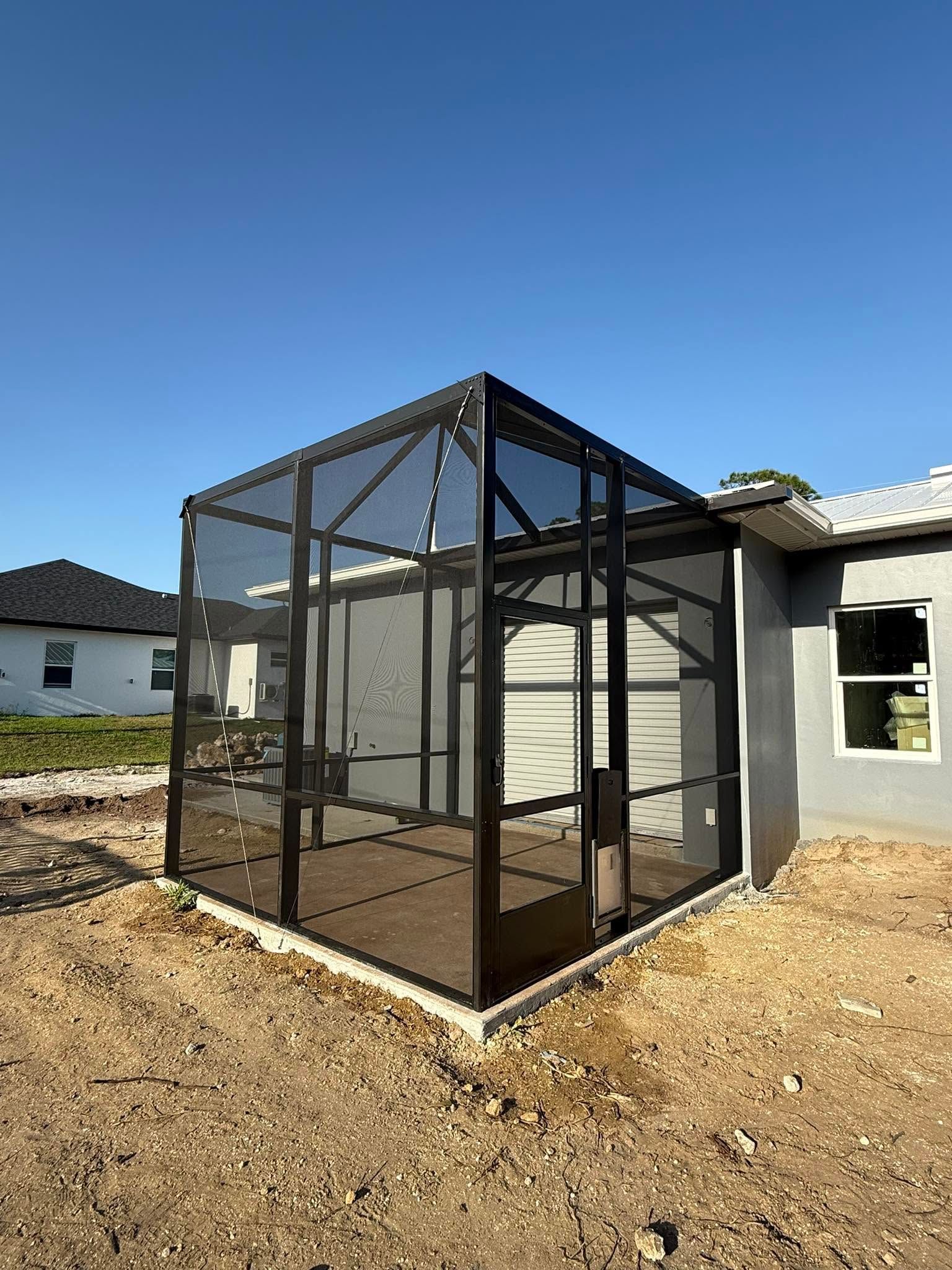 Black mesh-enclosed outdoor patio or screen room beside a house on a dirt lot under a clear blue sky