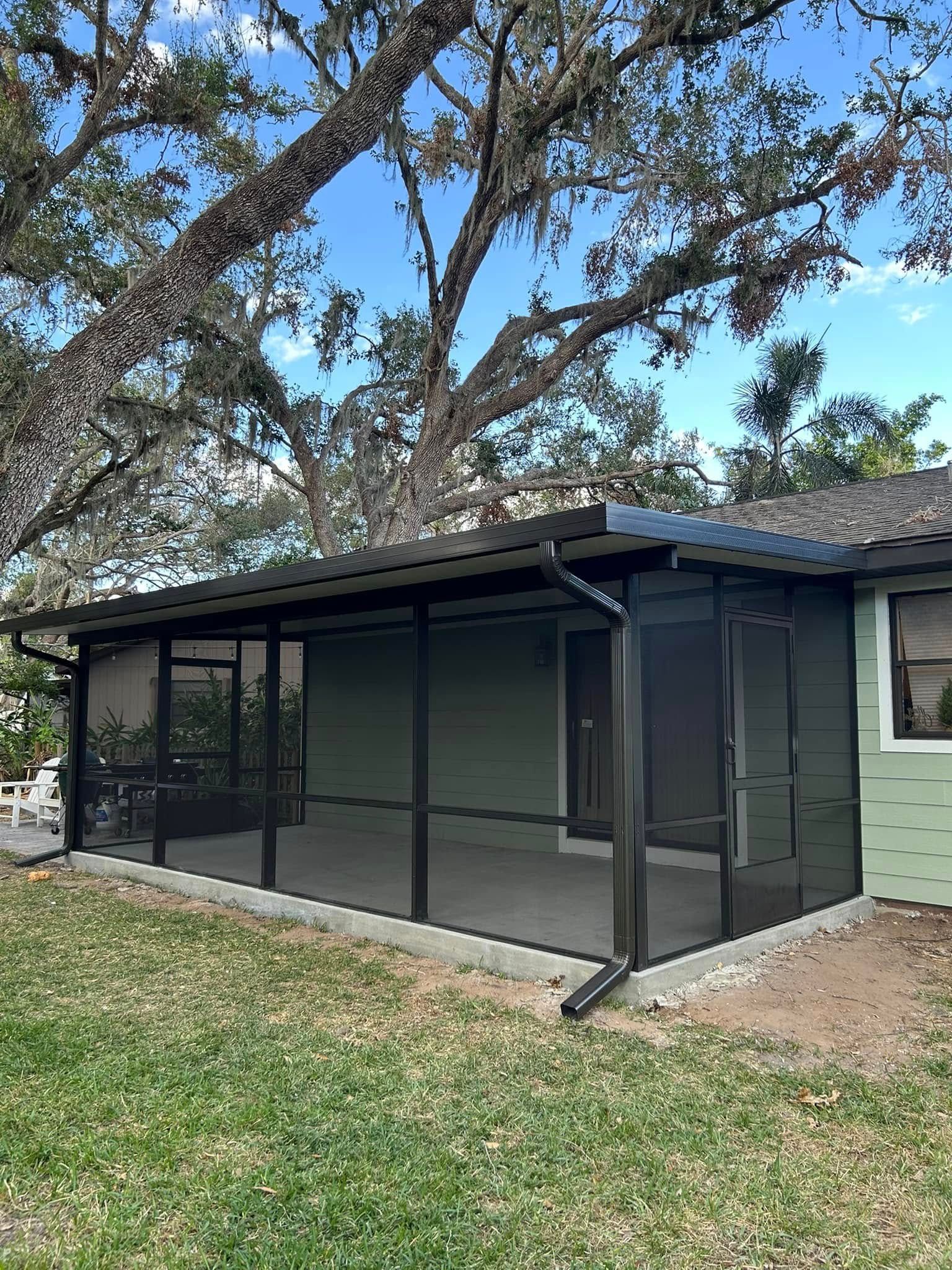 Screened porch with black framing attached to a light green house under large oak trees