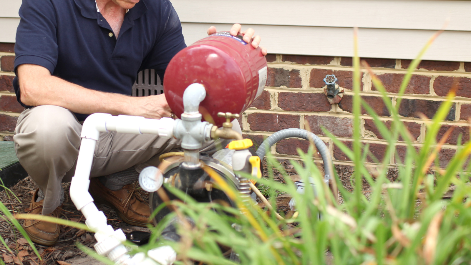 Person crouches near a red fire hydrant by a brick wall, handling white pipes amid tall grass.