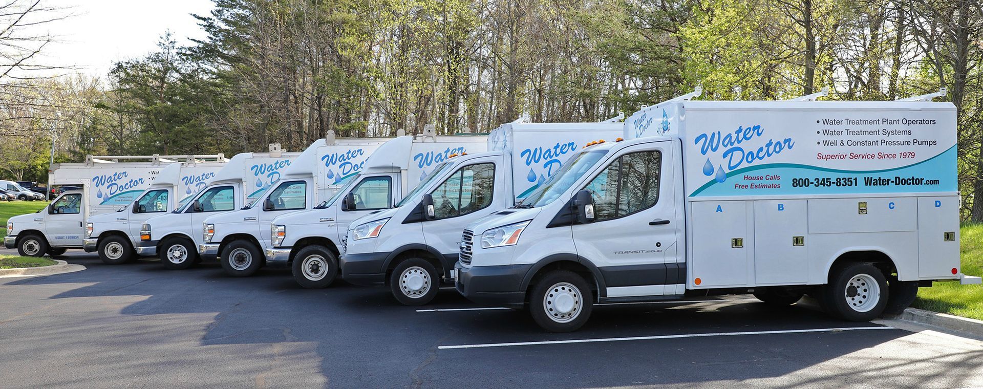Several white utility vans lined up in a parking lot under trees