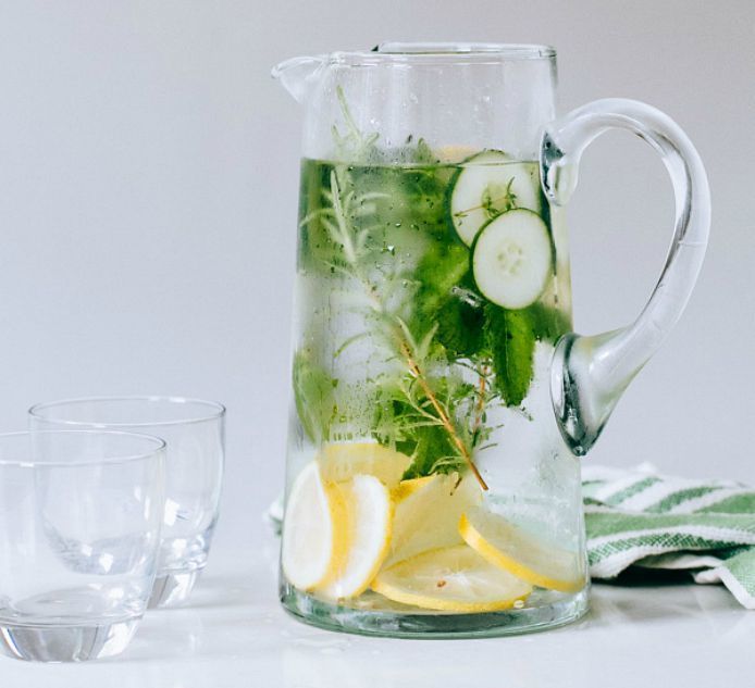 Glass pitcher of cucumber, lemon, and herb infused water beside a drinking glass and striped cloth.