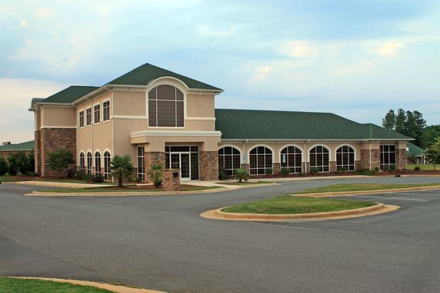 Large tan commercial building with green roof and arched windows beside an empty parking lot.