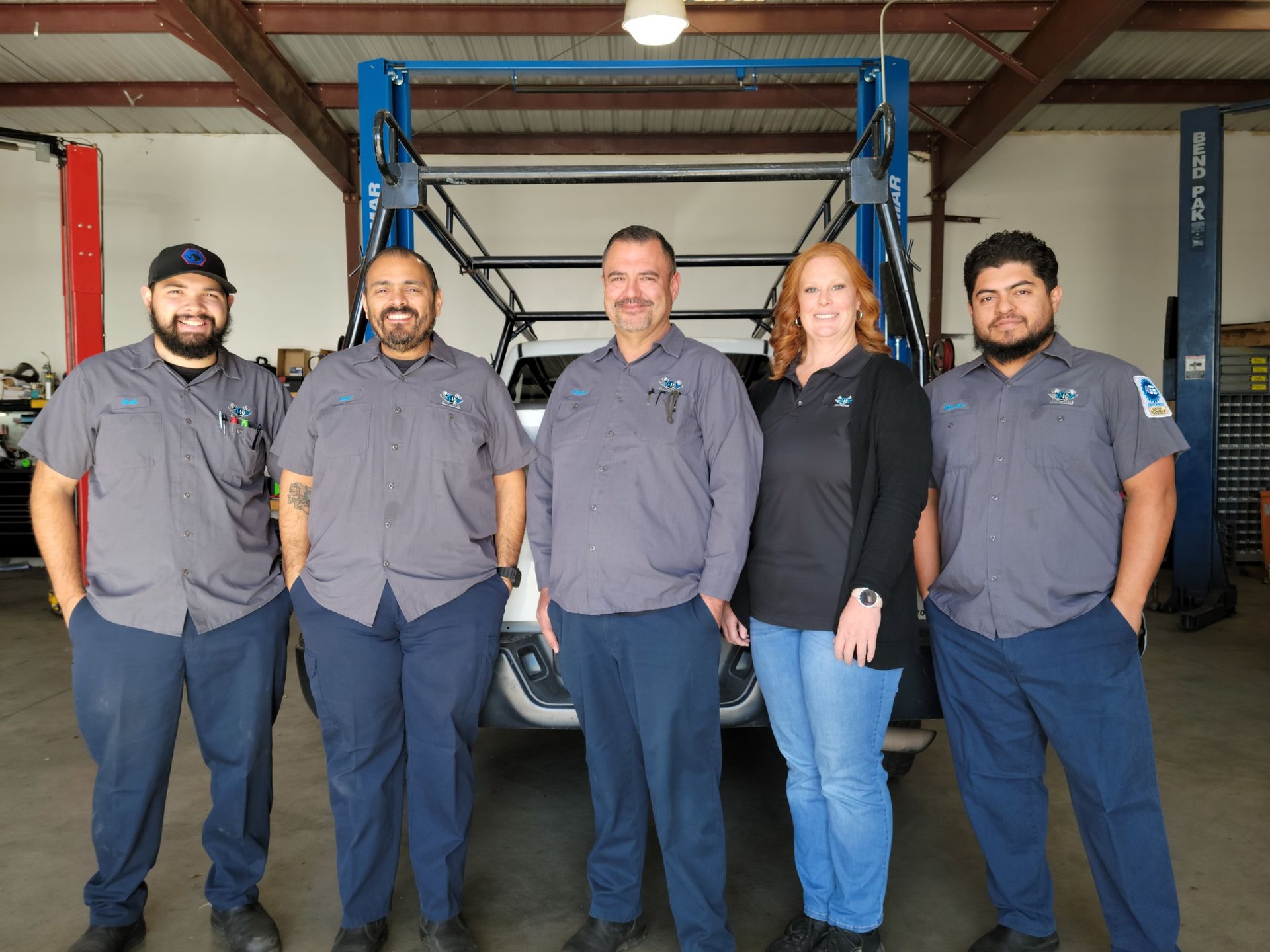 Five people in work uniforms posing in front of a vehicle lift in a garage.