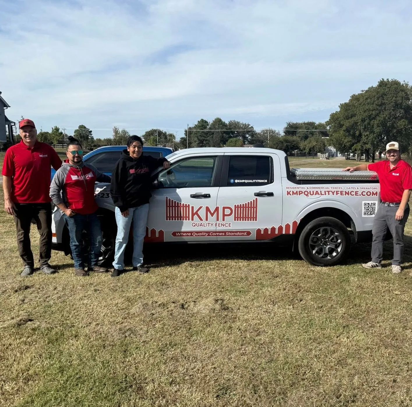 Four people stand beside a company truck on a grassy field under a blue sky. The truck is white with company logo.