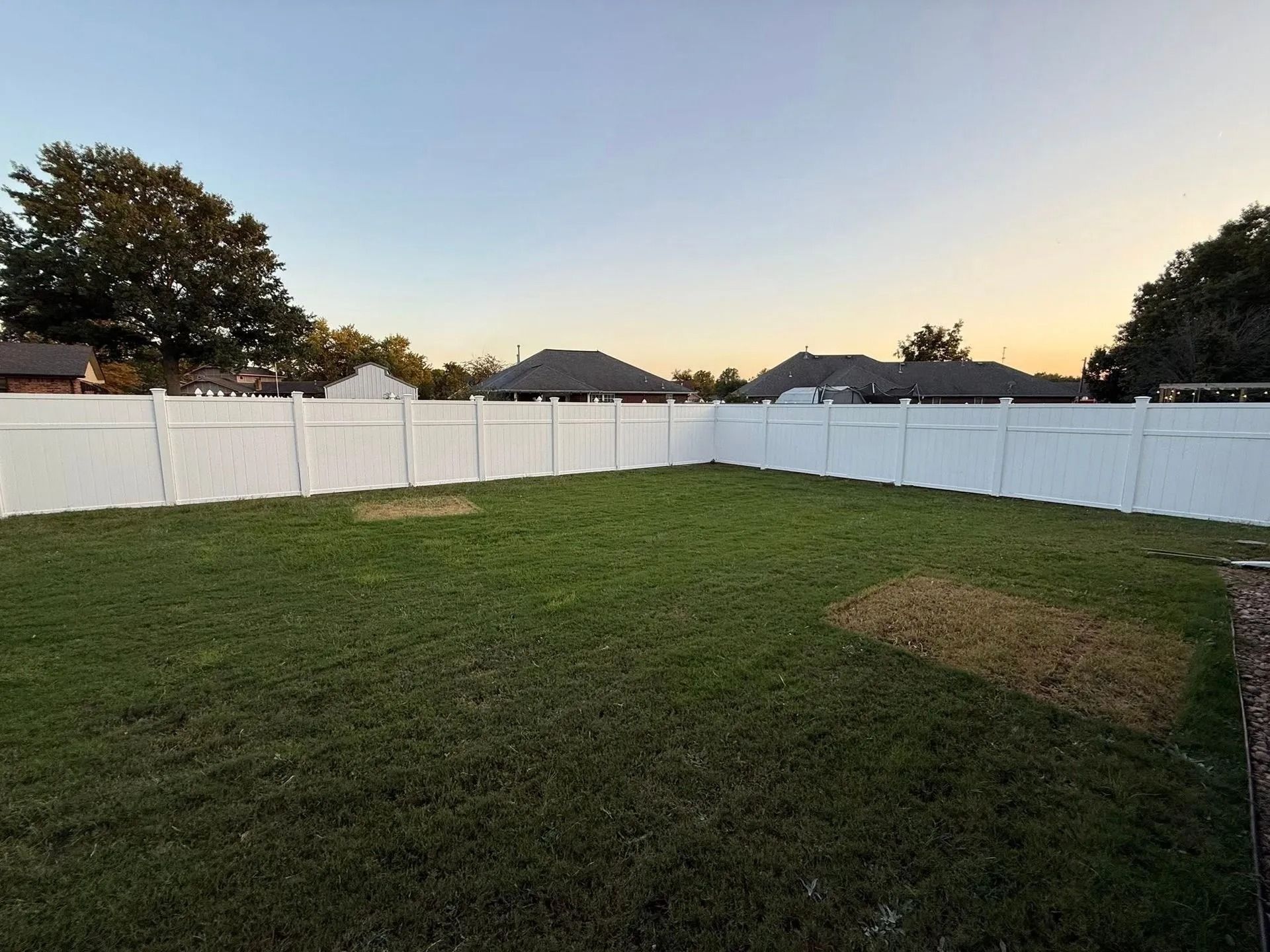 A white fenced backyard with green grass and a few brown spots, houses in the distance, and a blue sky.