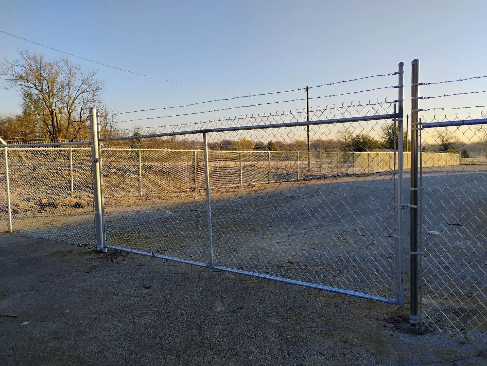 Chain-link fence with barbed wire on top, enclosing a gravel area, under a blue sky.