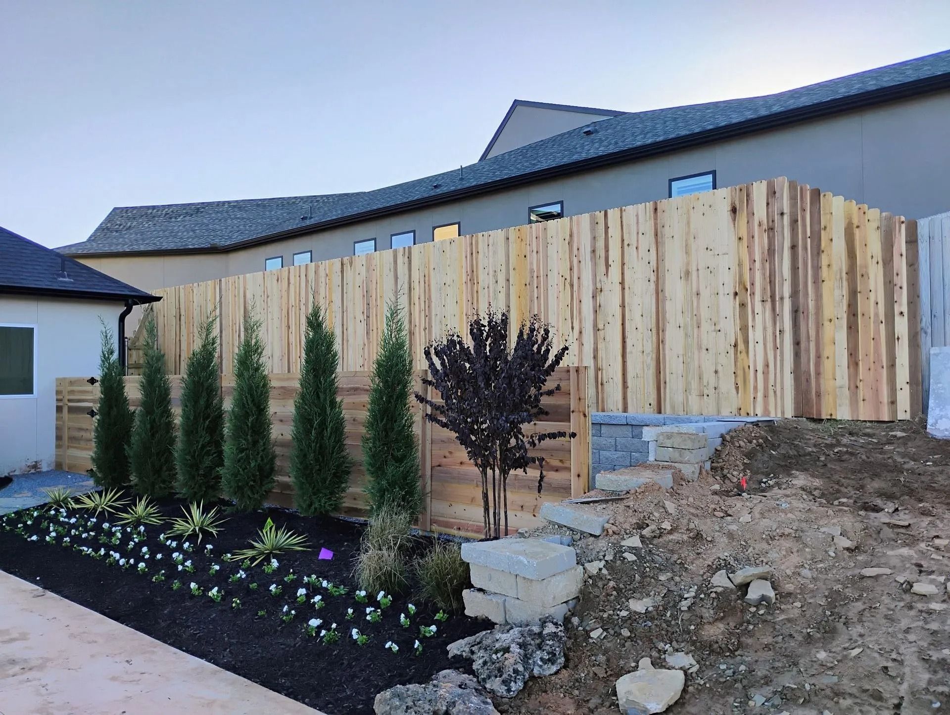Wooden fence bordering a landscaped garden with small evergreens and black mulch, next to a home.
