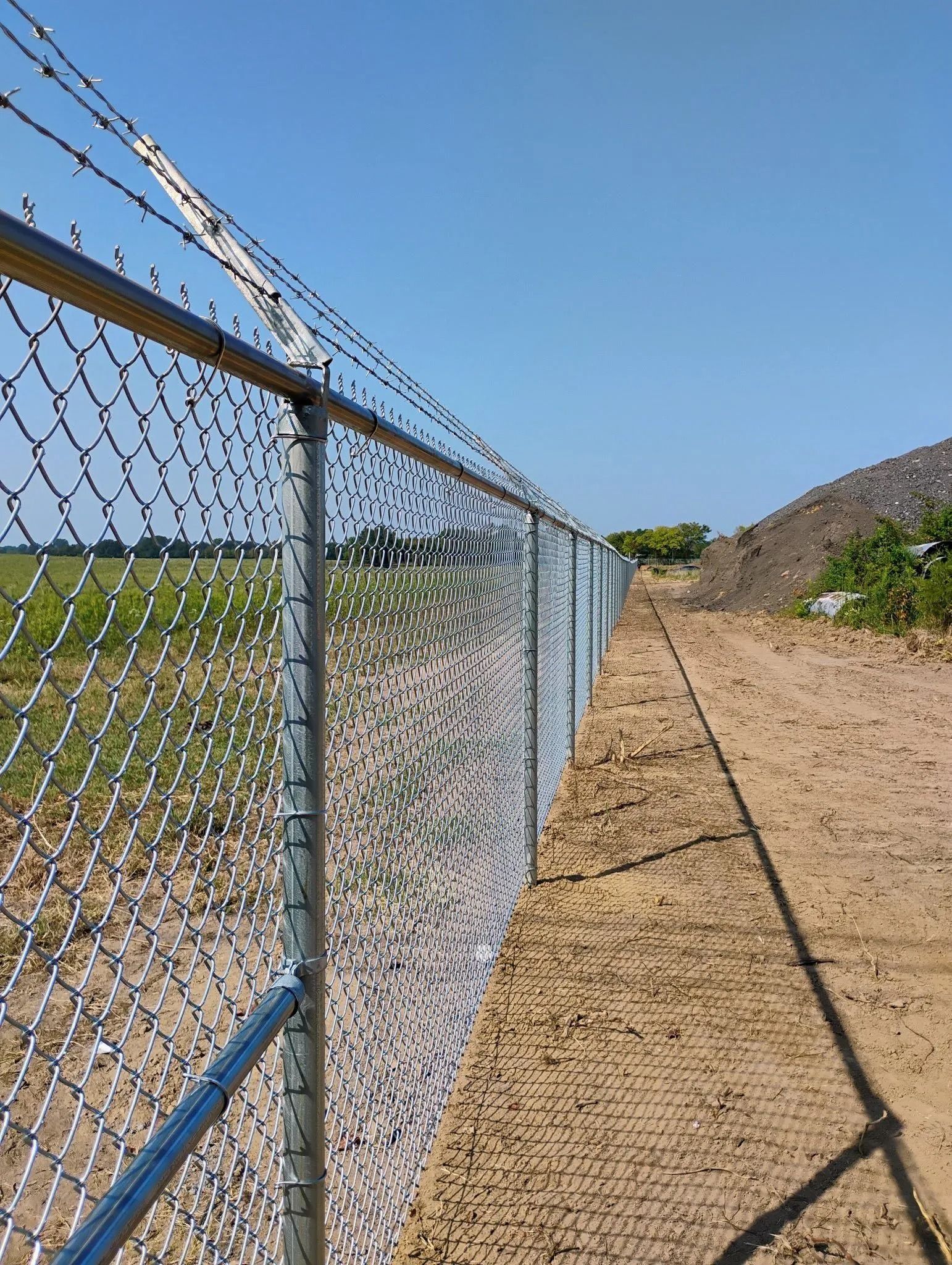 Chain link fence topped with barbed wire, alongside a dirt path under a clear blue sky.