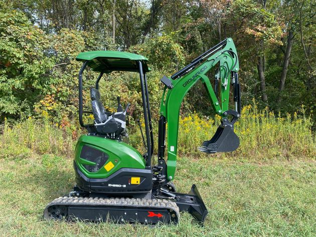 A small green mini excavator with a black seat and rubber tracks, parked on a grassy field in front of green trees.